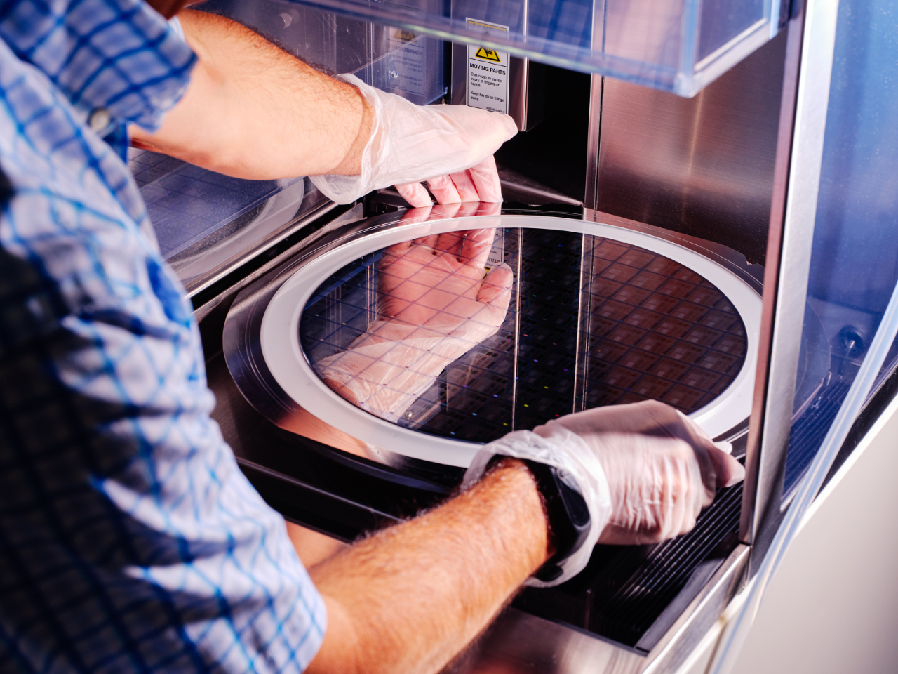 A technician carefully handles a large circular semiconductor wafer inside a specialized machine. The person wears disposable gloves and a short-sleeve checkered shirt while working with the equipment. The wafer surface shows a grid pattern of microchips reflecting light. The scene suggests a cleanroom or laboratory environment focused on electronics manufacturing and precision technology.